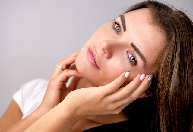 A woman with long brown hair and blue eyes gently touches her face with both hands against a neutral background. Her expression is calm, and she is wearing a white top.