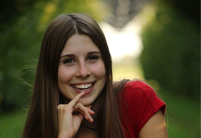 A woman with long brown hair and blue eyes gently touches her face with both hands against a neutral background. Her expression is calm, and she is wearing a white top.